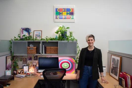 A young woman wearing a black blazer stands in an office cubicle space with artwork on the desk and one larger print framed above the desk. 