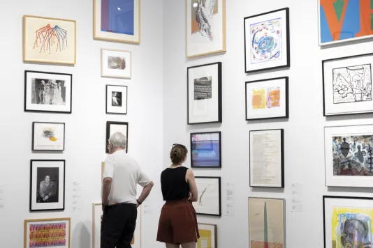 Gallery walls lined with framed artworks and two people facing the artworks viewing. 